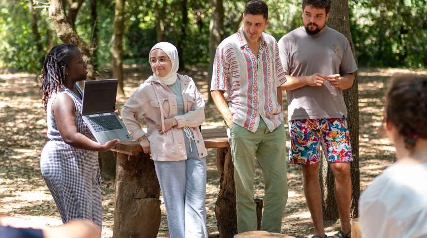 Four diverse adults in their 20s stand in a forest clearing presenting a project to peers who are seated nearby