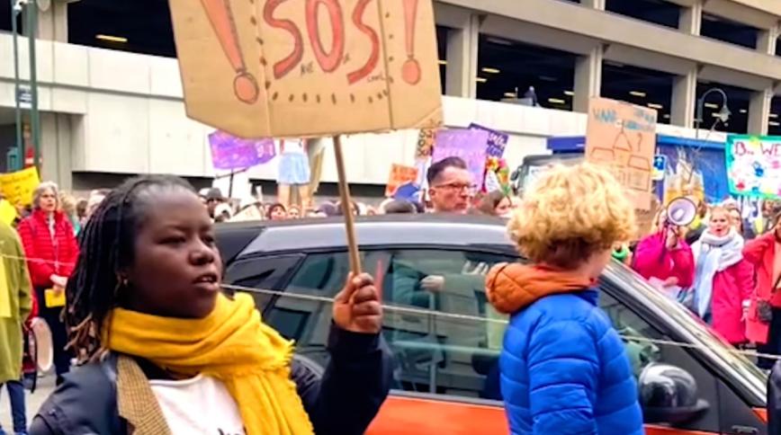 A Black woman with a yellow scarf around her neck holds a sign that says SOS in what appears to be a protest in the city streets