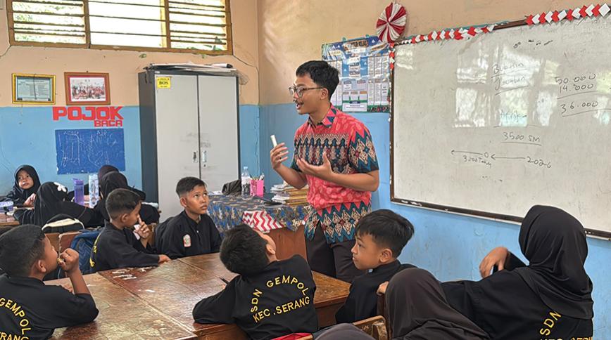 A teacher standing in front of a whiteboard, gesturing while presenting a lesson to students seated around tables in a classroom