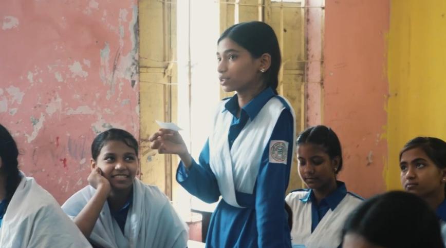 A young femle student with tan skin and long brown braids wearing a blue and white school uniform stands in a classroom holding a paper to speak while her classmates listen
