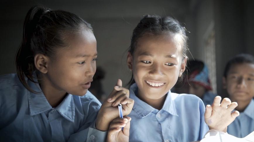 Two Asian girls wearing blue blouses smile while one holds a pen