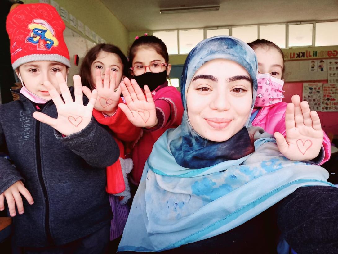A smiling young woman wearing a blue and white head scarf is surrounded by very young girls all holding up their hands on which hearts are drawn