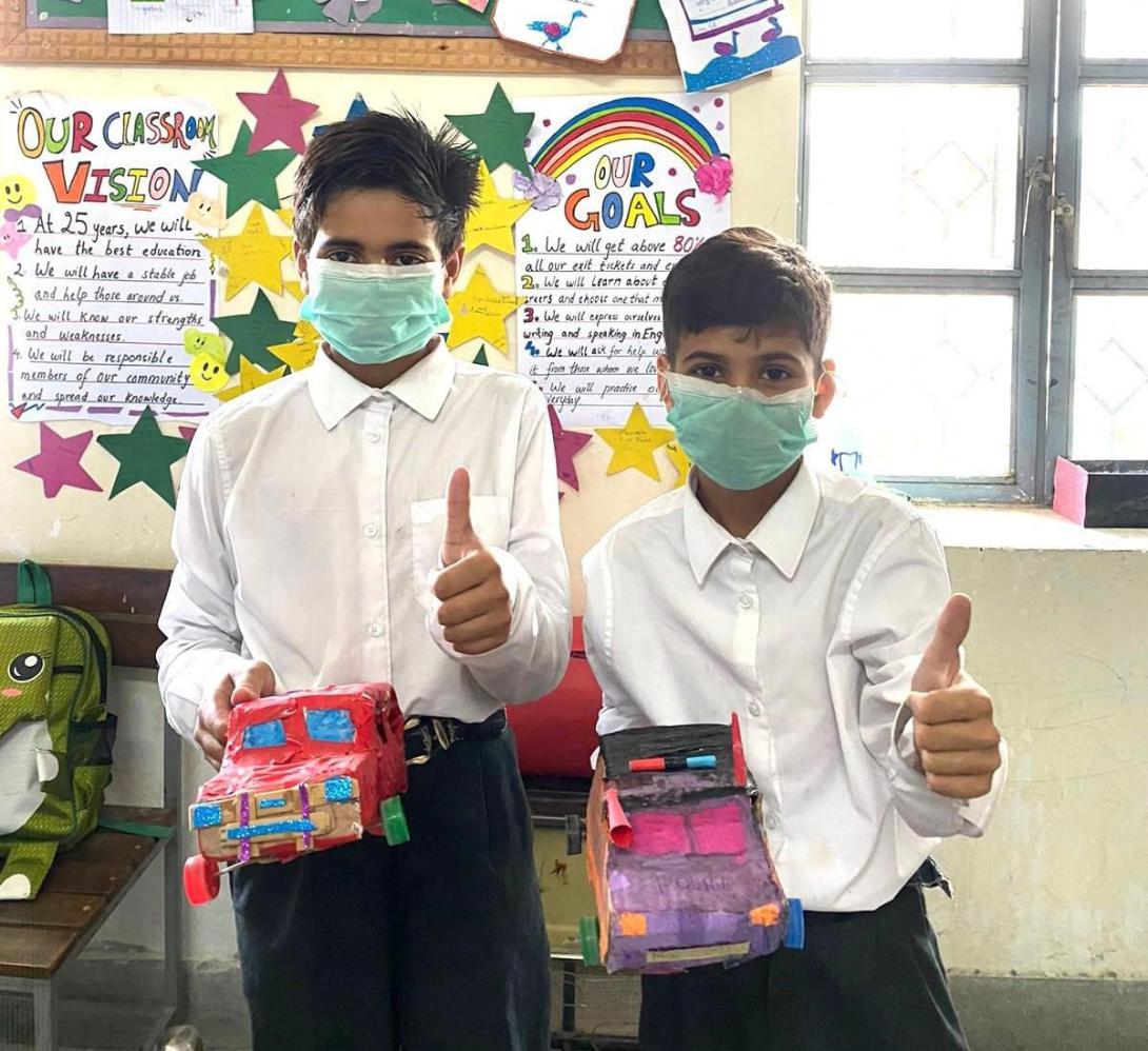 Two boys with brown skin and dark hair hold vehicles made out of cardboard and give the thumbs-up sign