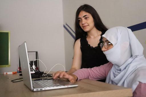 A young female teacher watches as a teenage girl wearing glasses and a headscarf types on a computer