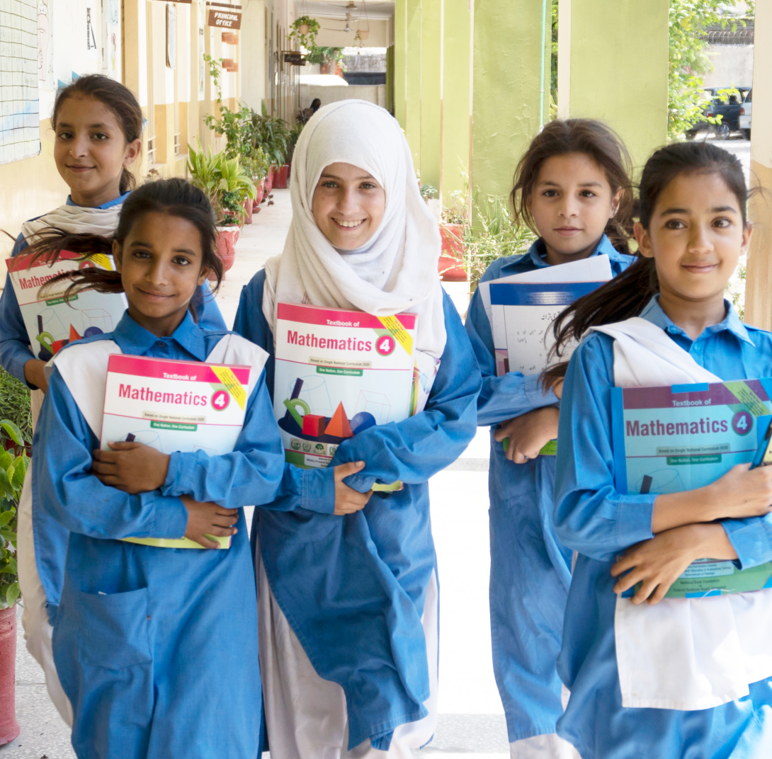 Five girls in blue dresses, one wearing a white head scarf, face the camera clutching mathematics workbooks
