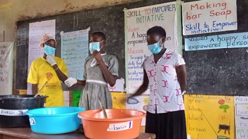 Three women mix materials in plastic basins in front of a chalkboard