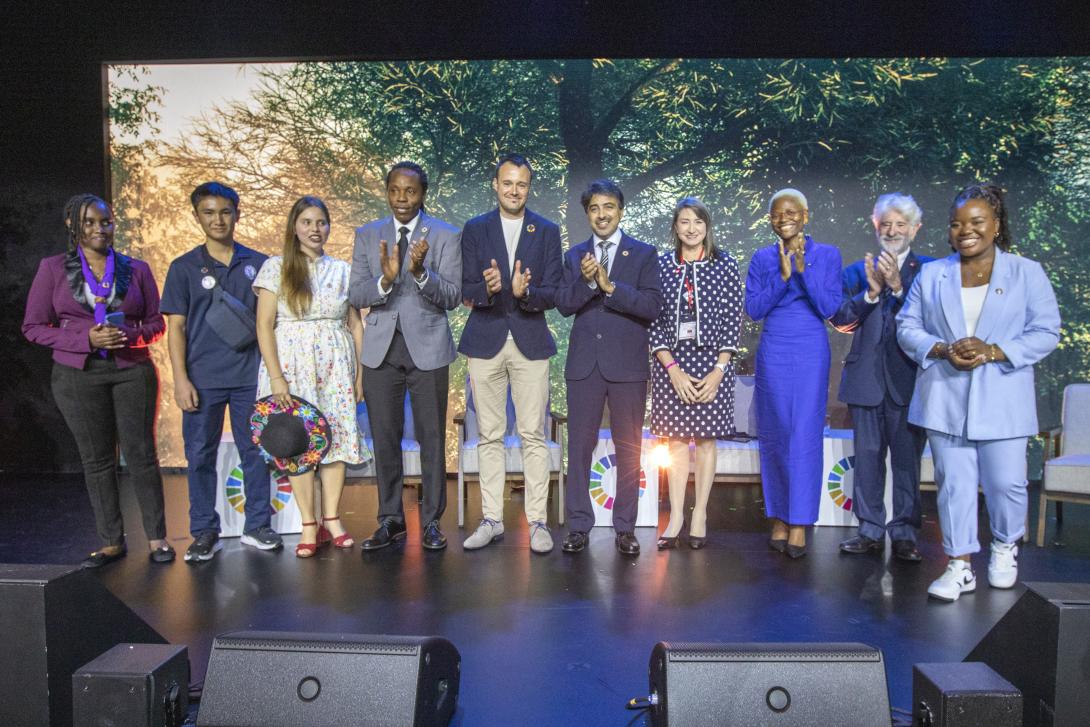 A row of 10 adults of diverse ethnicities stands on a stage clapping in front of an image of leafy treetops