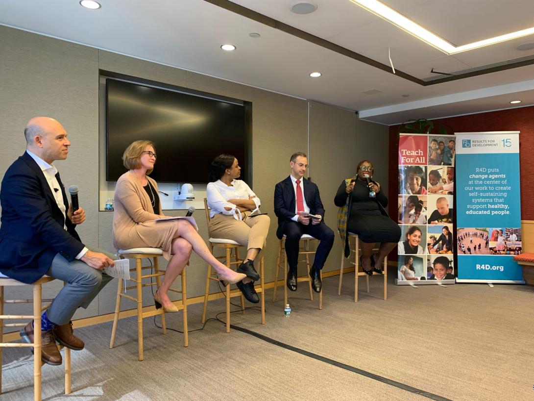 Five people sit in a row in the front of a room. A Black woman on the far right speaks into a microphone