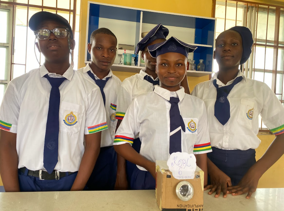Four Nigerian teens in school uniforms stand behind a camera made out of a cardboard box and other materials