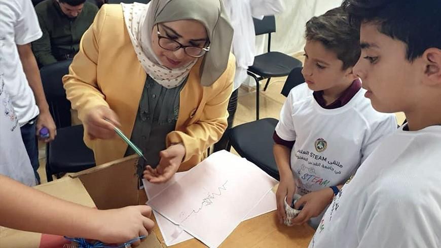 A female teacher wearing a head scarf demonstrates a science activity to two boys and other children who are mostly out of frame