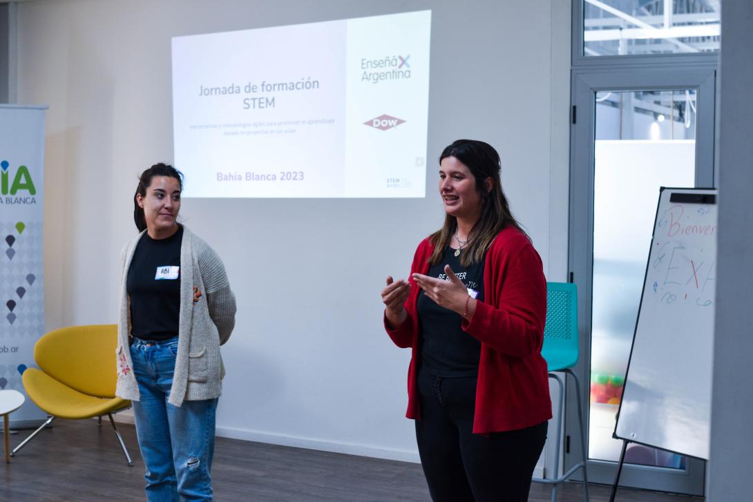 Two young women speak in front of a wall with a projection on it that says "Jornada de Formacion STEM" and includes DOW and Ensena por Argentina logos