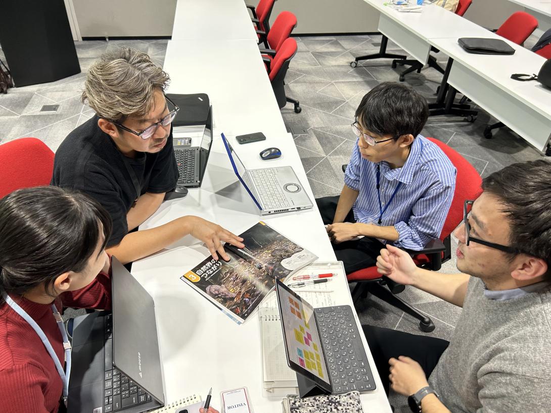 Three Asian men and one Asian woman sit at a table with their laptops while looking at a children's science magazine