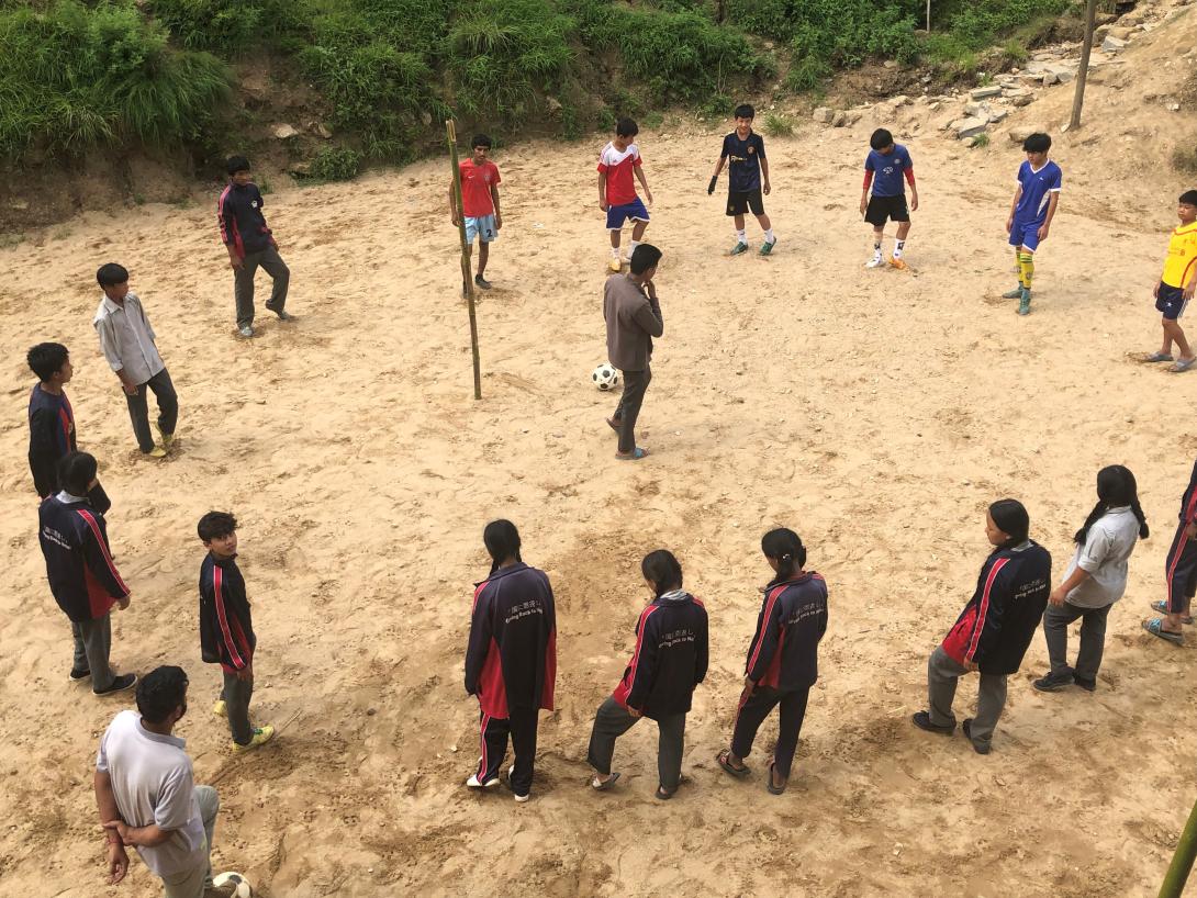Nepalese children stand outside in a circle around a teacher with a football