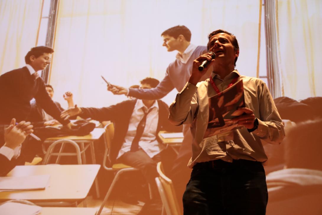 A man speaks into a microphone with an image of a teacher in a classroom behind him