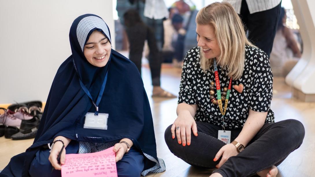 A young student in a hijab sits on the floor next to a blonde, white woman. They are smiling and talking