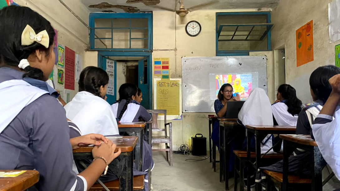 Students seated at desks in a classroom, shot from behind their heads as they look at a female teacher presenting in front of the whiteboard