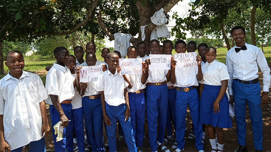 A large group of Nigerian students stand outside with their teacher. They are standing under a tree and holding handwritten signs with messages like "Dream big, work harder" and "Discover your career"