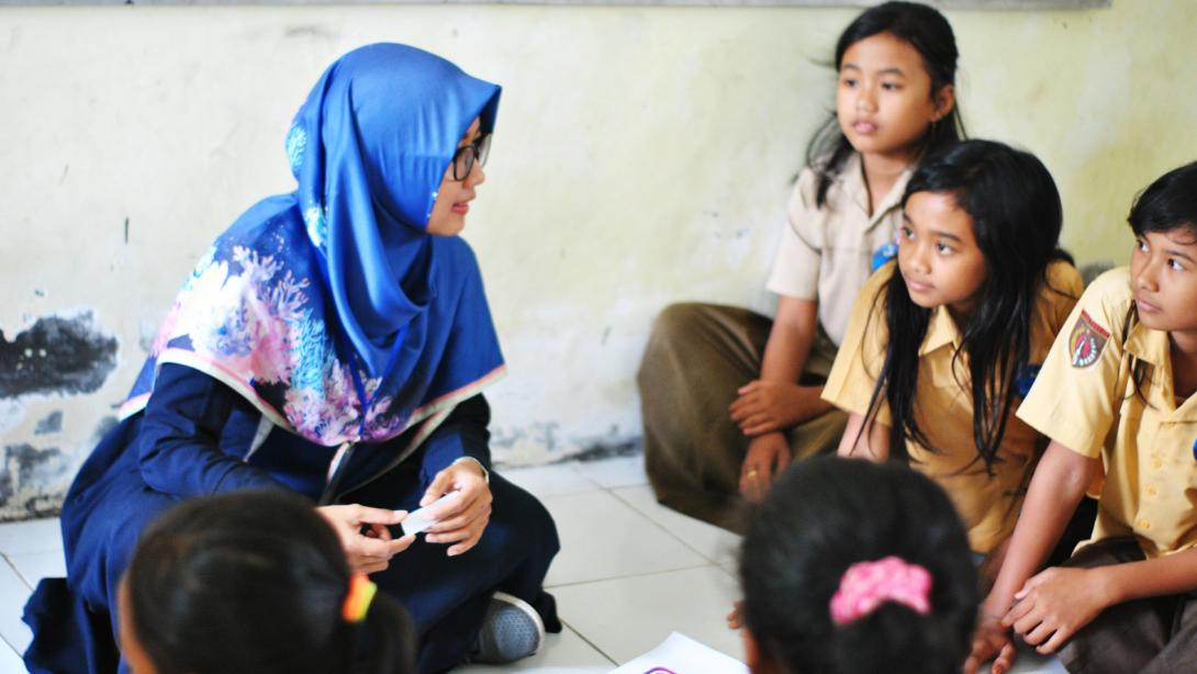 A teacher in a blue hijab sits on the floor talking to young students in their school uniforms