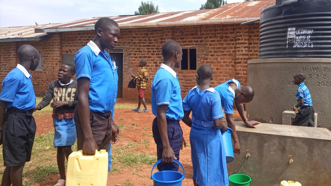 Ugandan students in school uniforms fill up water jugs at a water tank labeled "Ugandan Water Project"