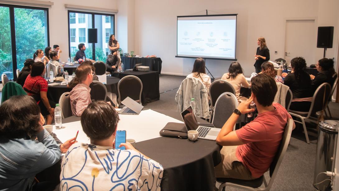 Room filled with people sitting in groups around tables and watching a presentation