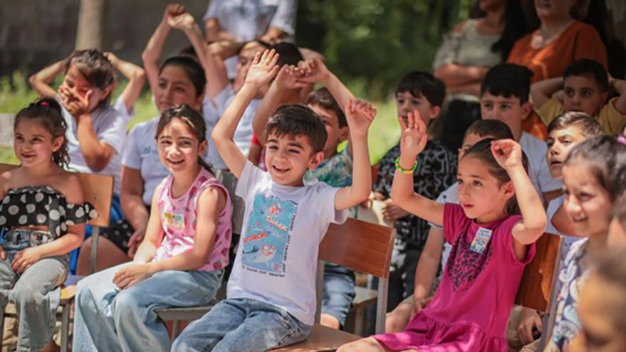 A group of children in colorful casual clothing sitting on chairs smile and some raise both hands above their heads