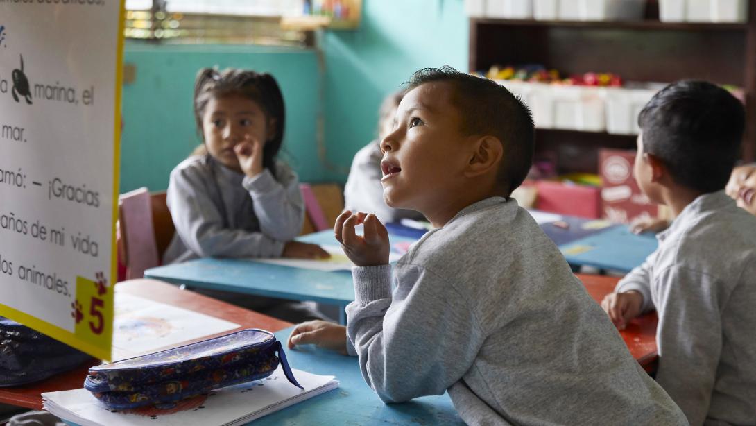 Four young children in school uniforms sit around a desk, the closest to the foreground is a young boy who is looking up at a chart