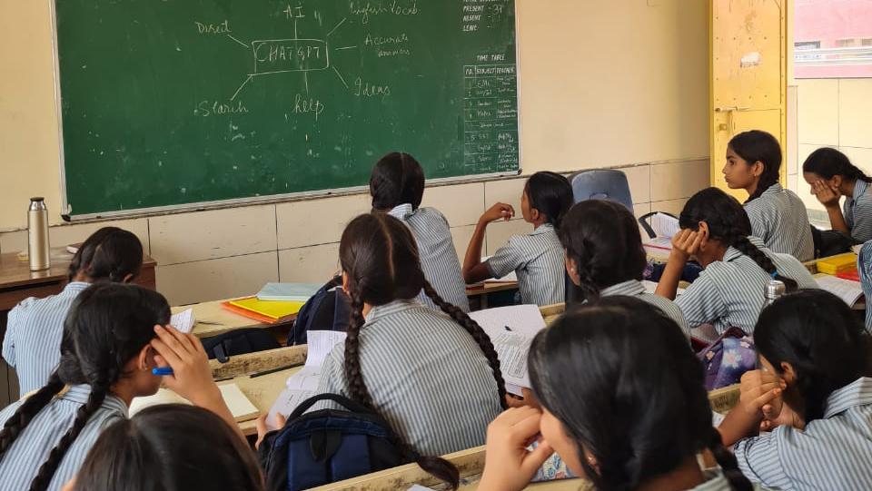 Young girls in school uniforms sit at desks looking at a chalkboard with a web diagram with "ChatGPT" in the middle written on the board