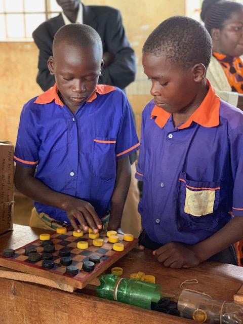 Two young Black boys in blue shirts with orange collars stand in front of a project involving a checker board, one of them touching one of the pieces