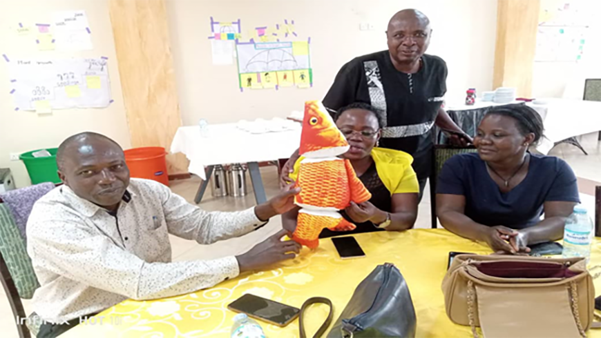 Four African adults sit at a table in the classroom. Two of them are holding up a bright orange fish icon