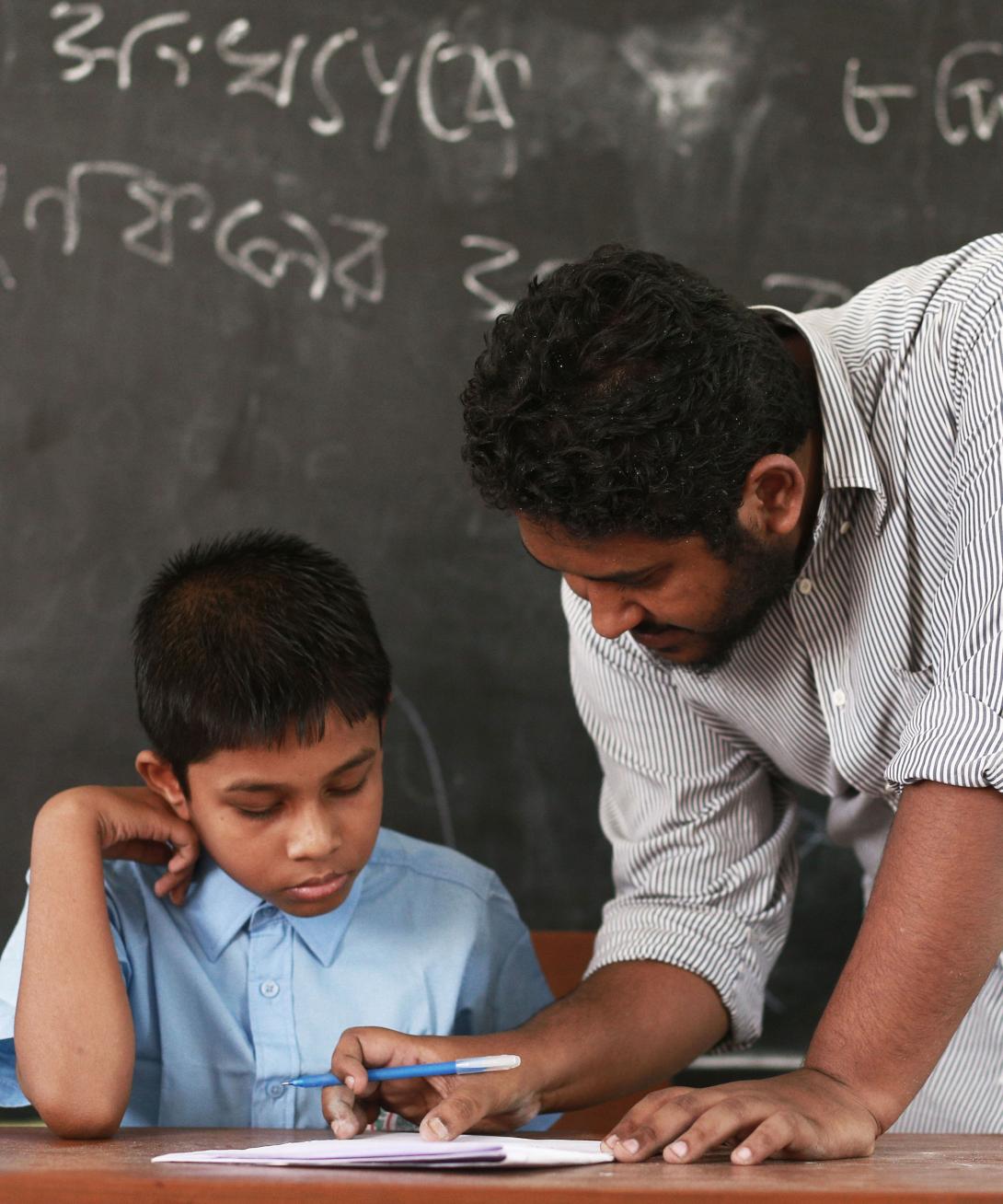 In front of a blackboard with writing on it, a male teacher with tan skin and brown hair leads over paper in front of a boy with similar coloring wearing a blue shirt