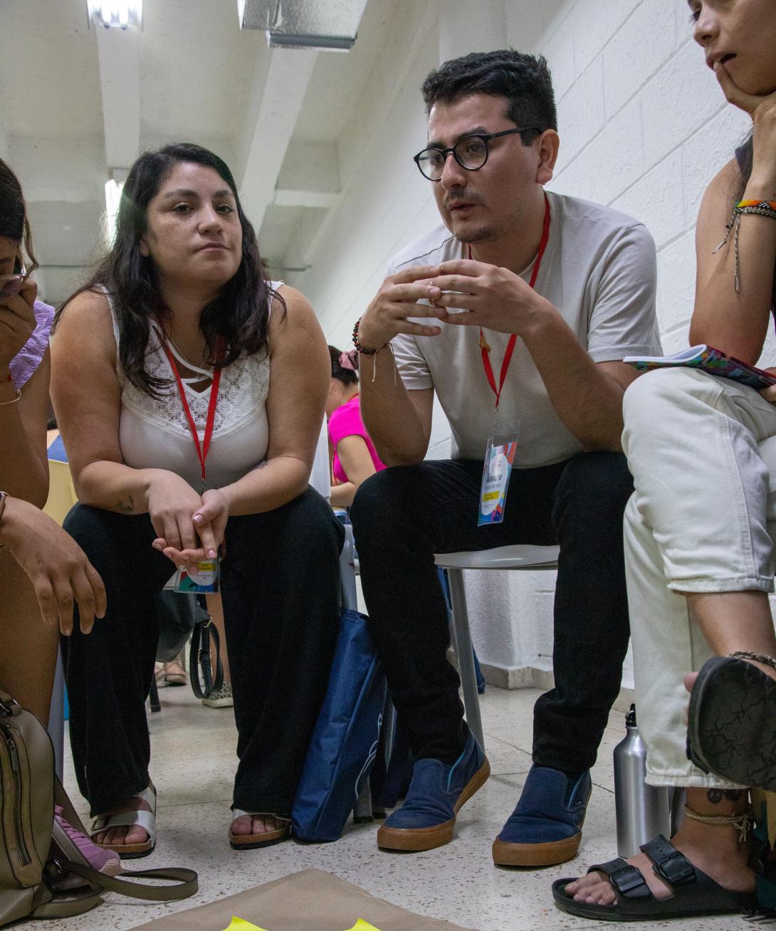 A young man and young woman sit in chairs among others surrounding a piece of paper on the floor with sticky papers on it