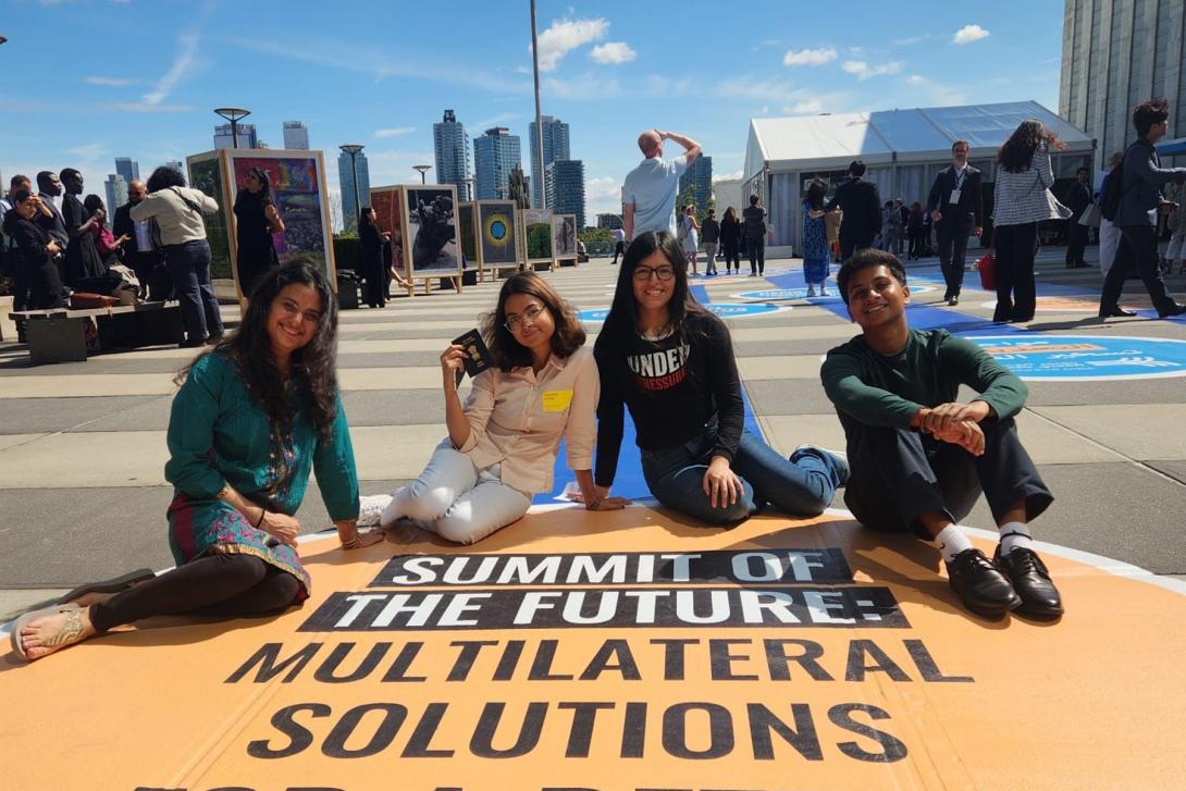 Four diverse young people sit on a yellow street mural that says Summit of the Future Multilateral Solutions with a blue sky and NYC skyline behind them