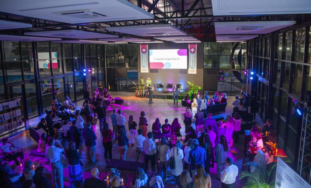 A crowd looks at a screen that says Ensena por Argentina in a large event space with many windows