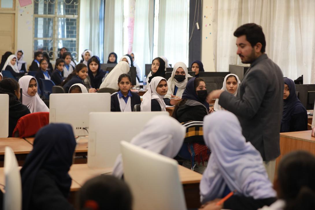 A man with dark hair and brown skin stands amidst a classroom full of girls in headscarfs sitting in front of computer screens and looking at him with attention