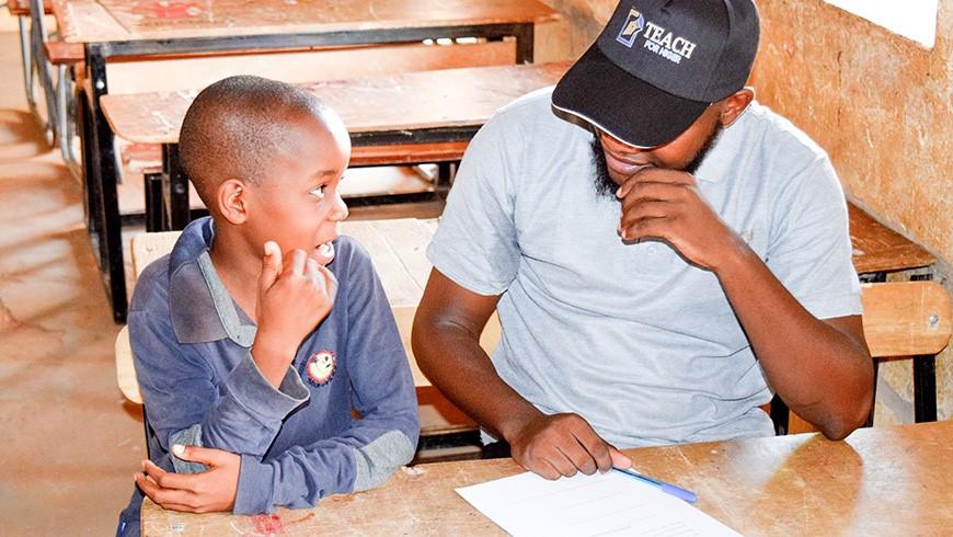 A young Black man in a baseball cap that says Teach For Niger and a young Black boy smile at each other while sharing a wooden desk in a classroom