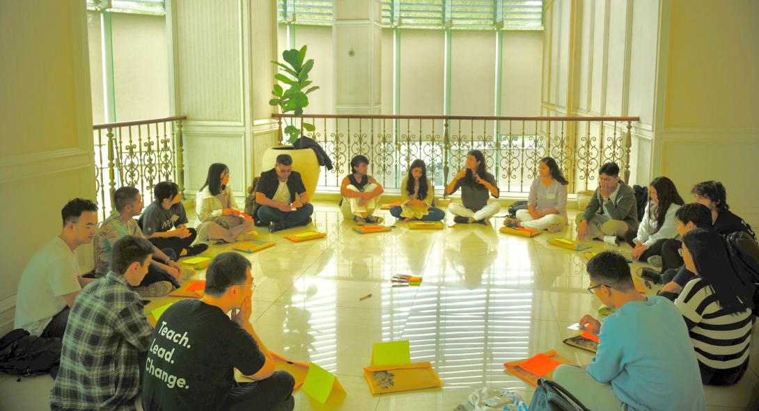 A diverse group of young people and young adults sit in a circle in front of a large window