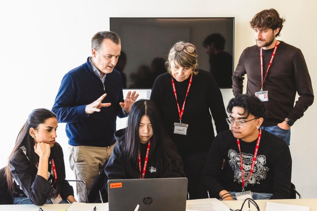 Three adults wearing name tags stand behind three teenage students, also wearing name tags, who surround a laptop