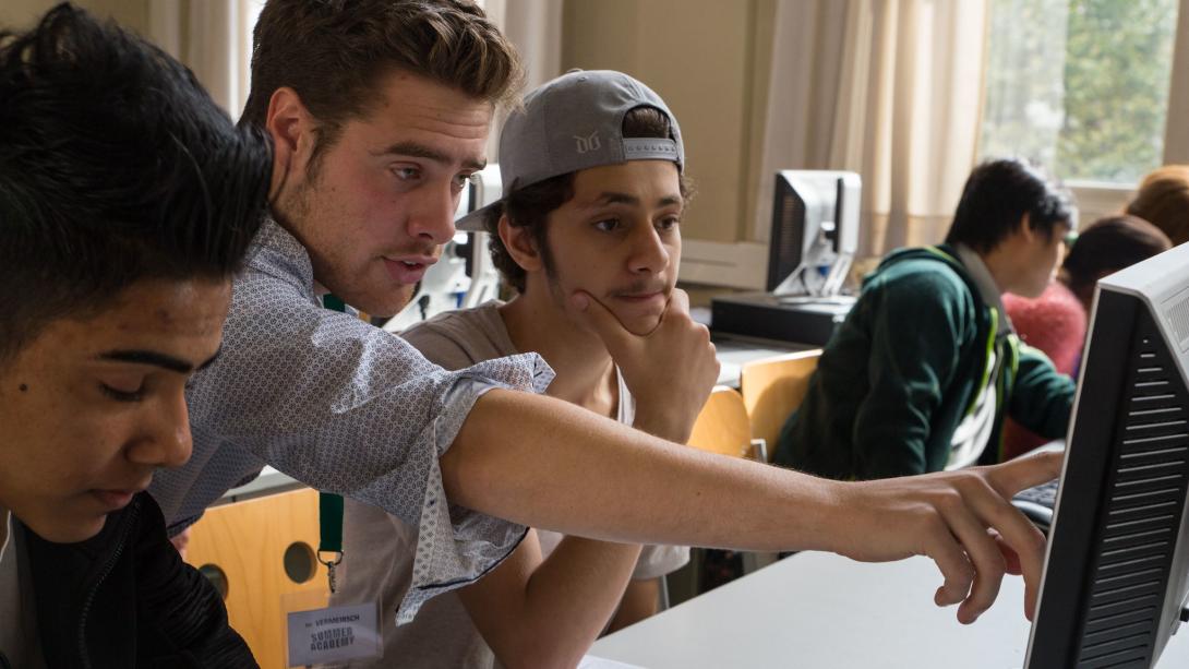 A young man points at a computer screen with teenage boys on either side of him, one in baseball cap