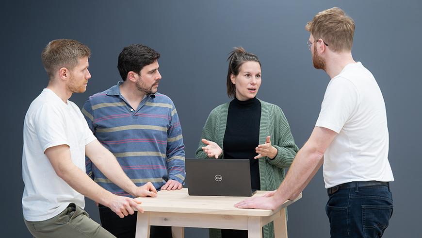 Three men and one woman stand around a table with a laptop on it. The woman is talking and gesturing with her hands and the men are all looking at her