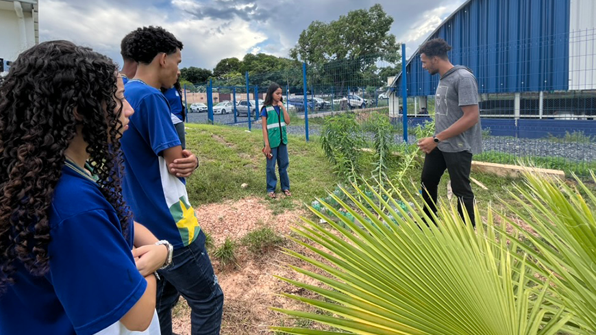 A group of students in blue uniforms stands in a grassy outdoor area, listening to a young man explain a gardening project involving small plants and plastic bottles