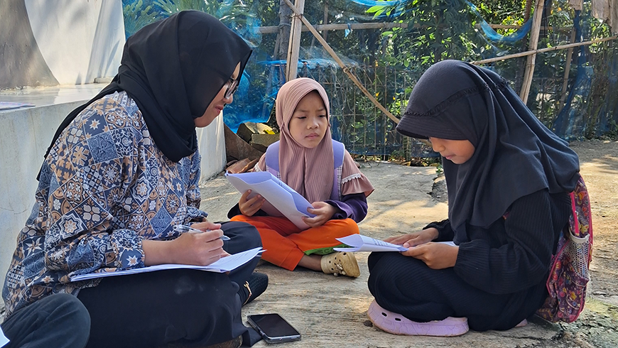 An educator wearing a black hijab and a patterned shirt sits on the ground outdoors, reviewing papers with two young schoolgirls who are also wearing hijabs.