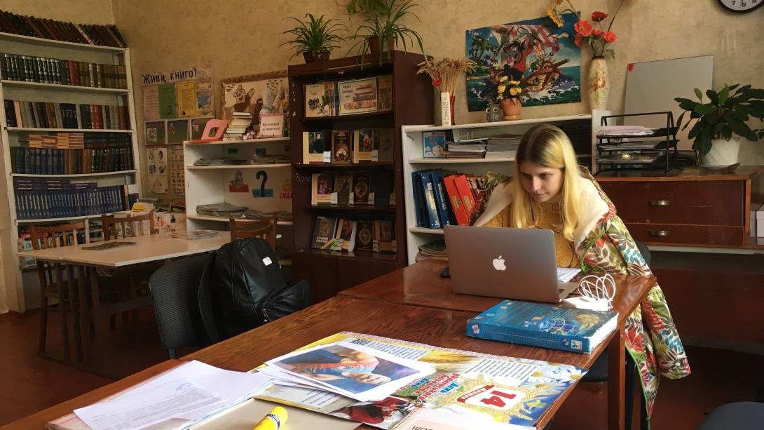 A young woman withe blonde hair and wrapped in a colorful blanket sits at a wooden table working on a laptop inside a classroom library filled with bookshelves.