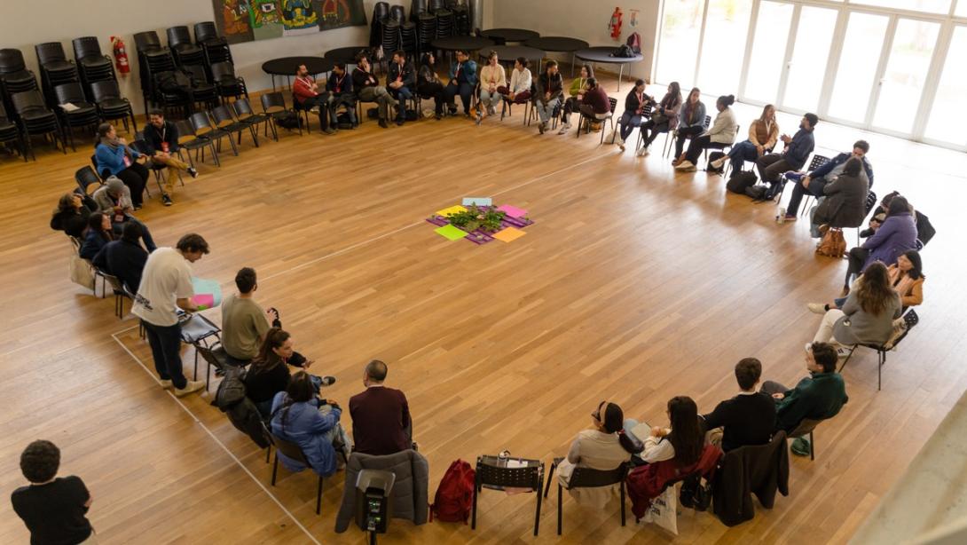 High-angle view of a large group of people seated in a wide circle on a wooden floor around a central display of colorful papers and greenery.