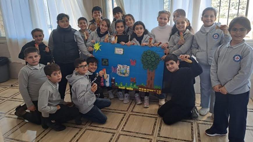A classroom group of school children in grey uniforms posing indoors around a large blue poster project showcasing nature themes, including a tree crafted with bottle caps, butterflies, flowers, and the Earth.